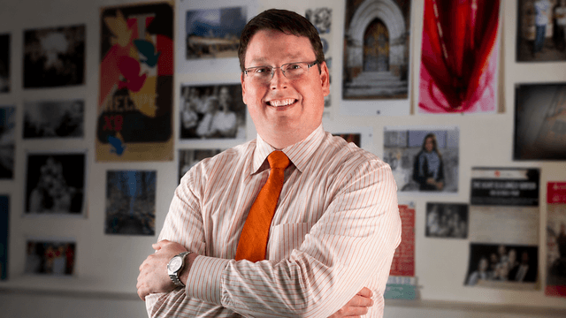 Nathan Hickman smiling with folded arms in front of a wall displaying photos and artwork.