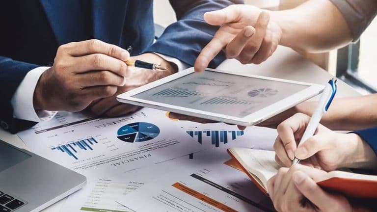 Three people in a business meeting reviewing charts on a tablet and printed documents. Hands are pointing and writing, conveying a collaborative focus.