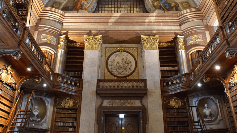 Historic library interior with ornate bookshelves, balconies, marble columns, and decorative ceiling artwork.