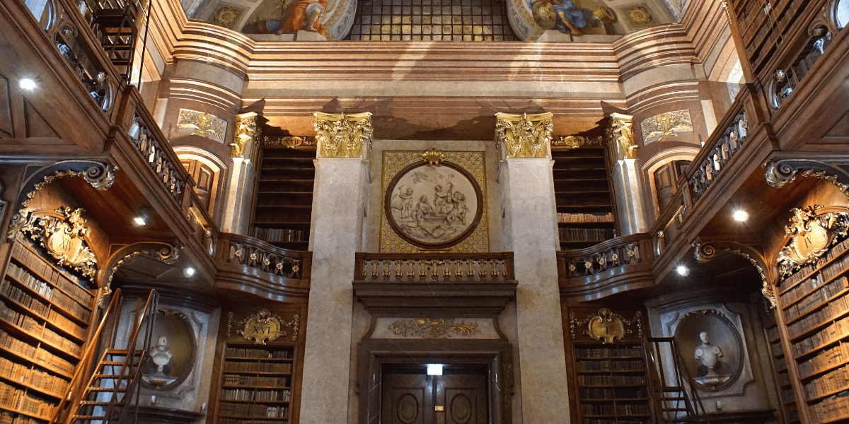 Historic library interior with ornate bookshelves, balconies, marble columns, and decorative ceiling artwork.