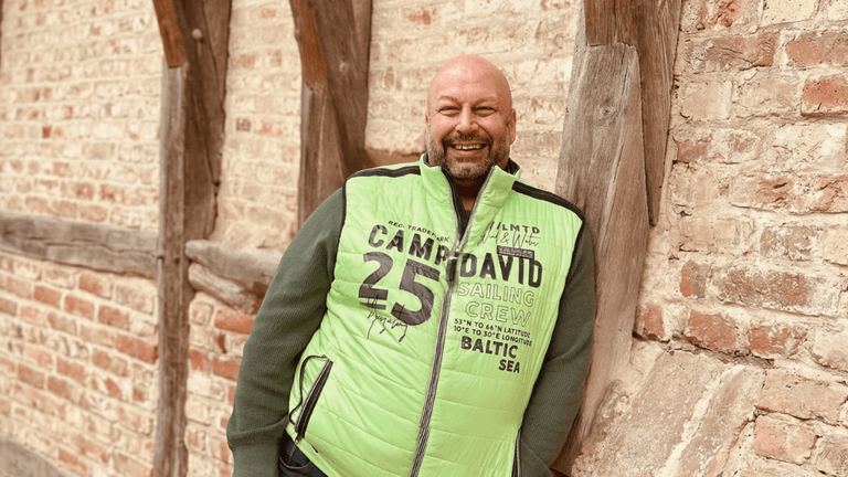 Heiko Gärtner smiling in a green vest, leaning against a rustic brick and timber wall outdoors.
