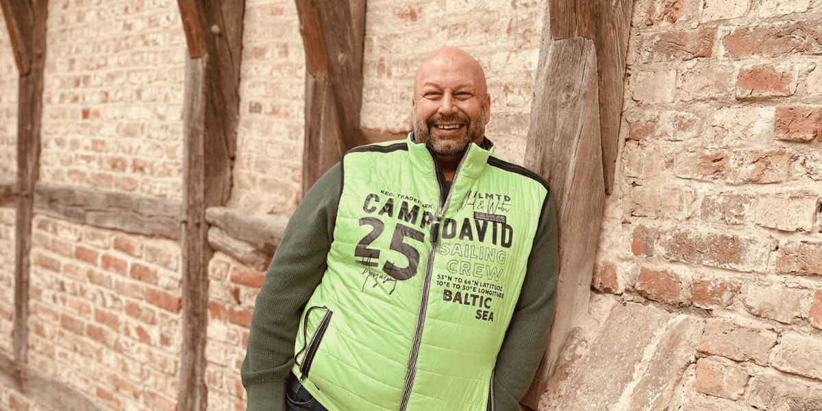 Heiko Gärtner smiling in a green vest, leaning against a rustic brick and timber wall outdoors.