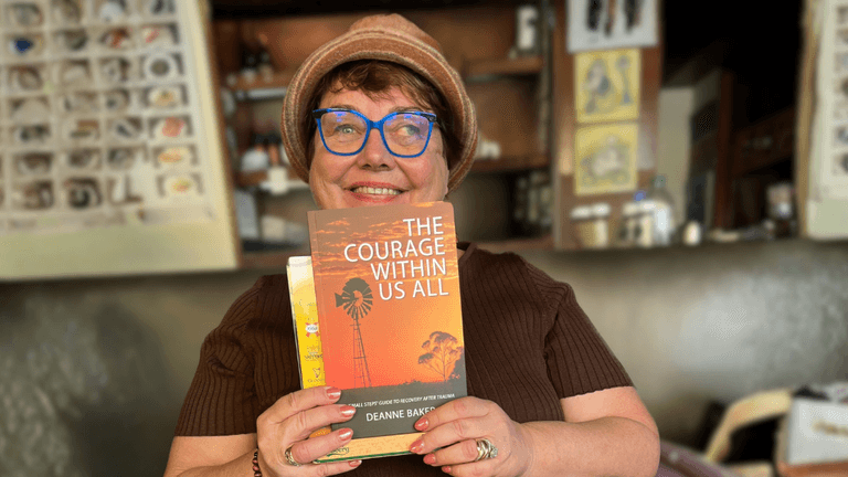 Smiling woman in hat and blue glasses holds memoir “The Courage Within Us All” inside cozy shop.