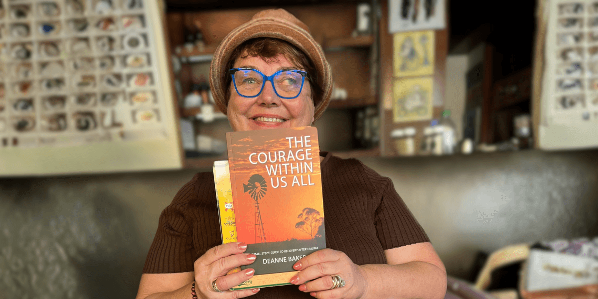 Smiling woman in hat and blue glasses holds memoir “The Courage Within Us All” inside cozy shop.
