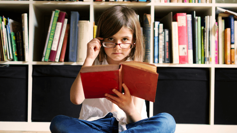Young girl sitting on the floor reading a book in front of a bookshelf
