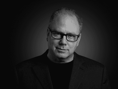 Man with glasses wearing a black shirt and blazer against a dark background