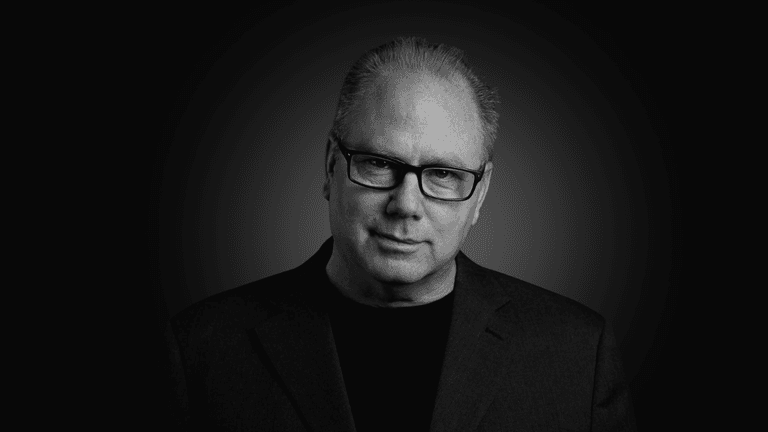 Man with glasses wearing a black shirt and blazer against a dark background