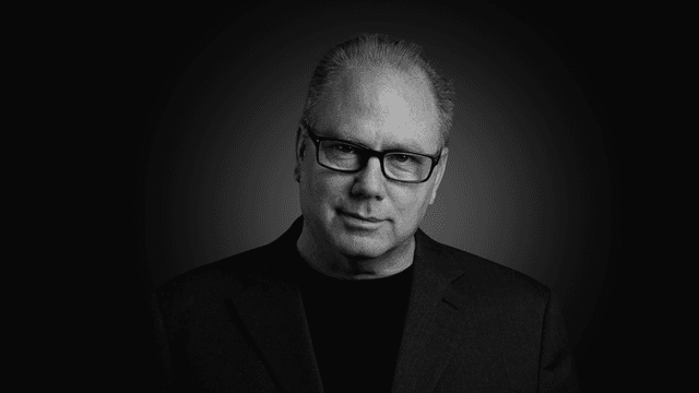 Man with glasses wearing a black shirt and blazer against a dark background