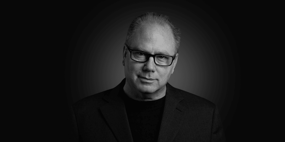 Man with glasses wearing a black shirt and blazer against a dark background
