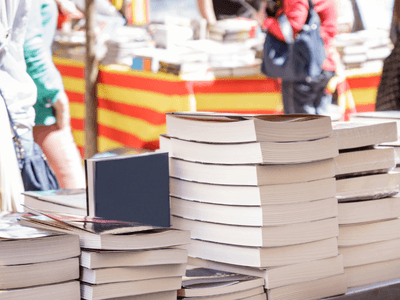 Stacks of books on a table with blurred vendors and colorful backgrounds in a market