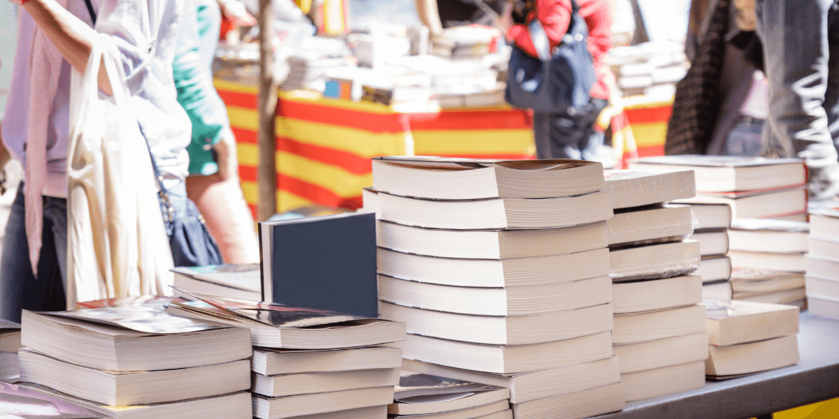 Stacks of books on a table with blurred vendors and colorful backgrounds in a market