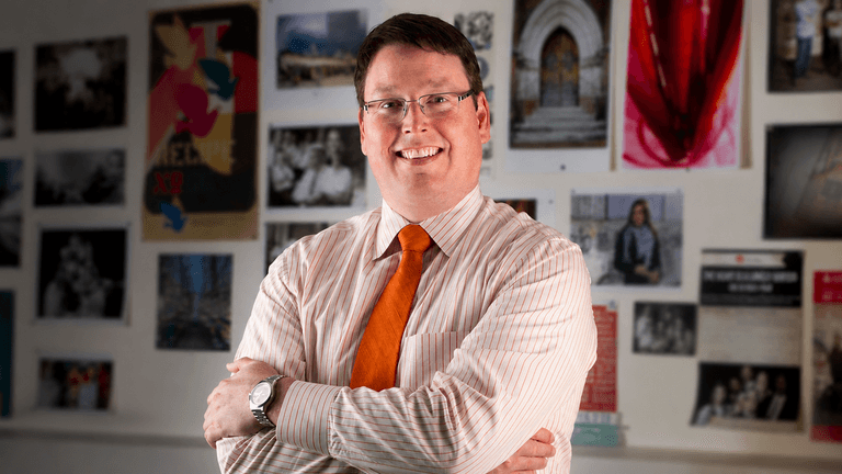 Nathan Hickman smiling with folded arms in front of a wall displaying photos and artwork.