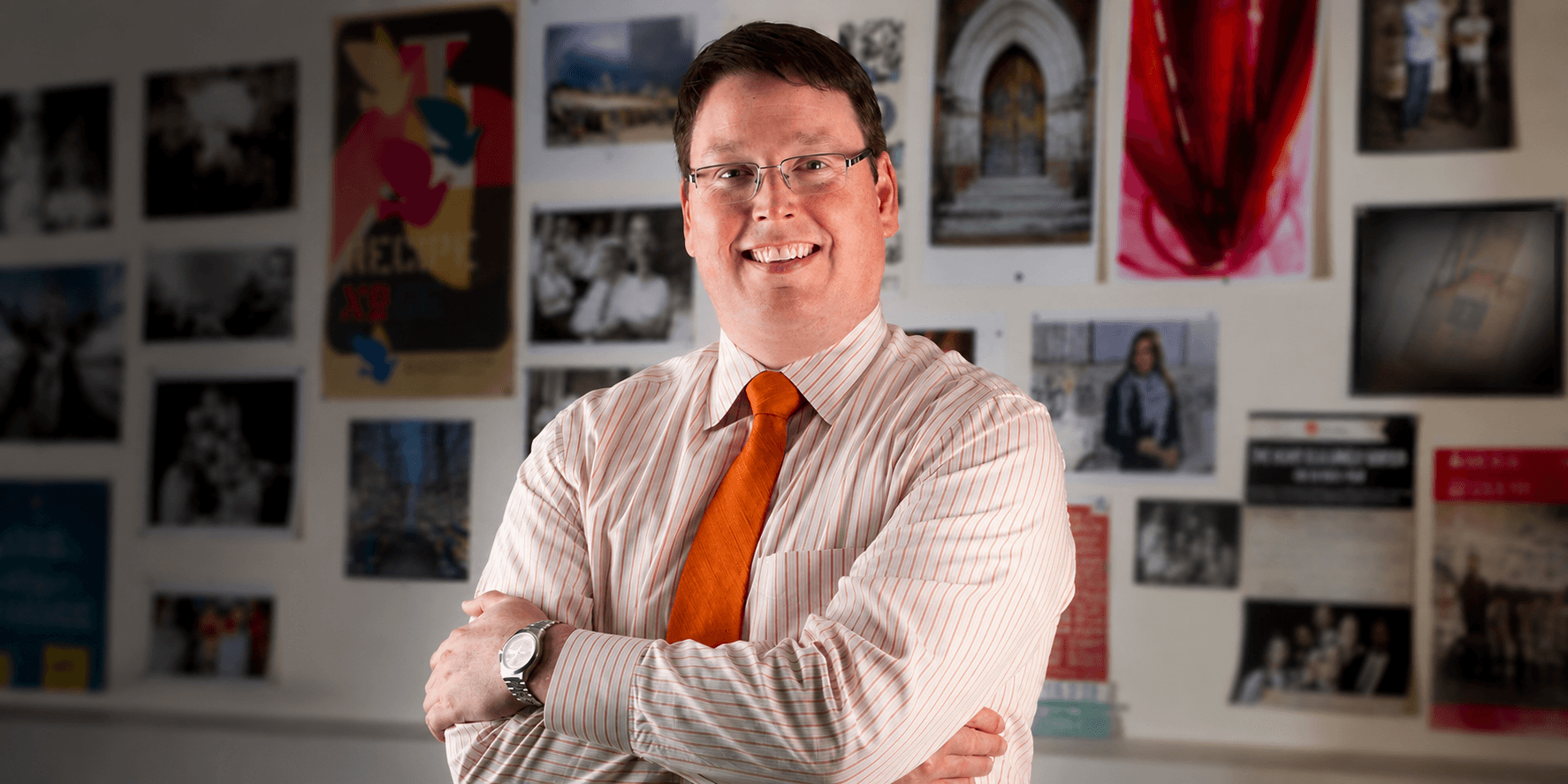 Nathan Hickman smiling with folded arms in front of a wall displaying photos and artwork.