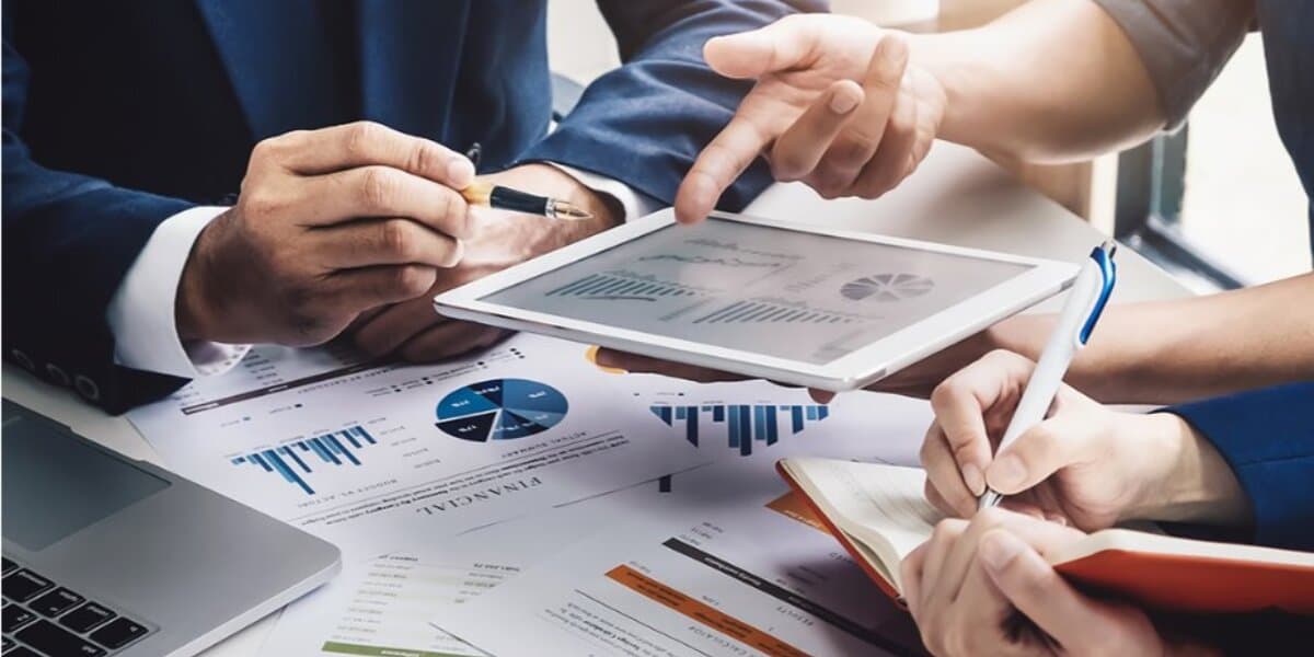 Three people in a business meeting reviewing charts on a tablet and printed documents. Hands are pointing and writing, conveying a collaborative focus.