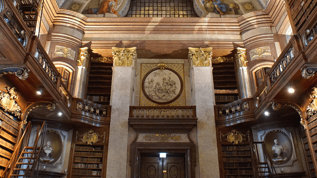 Historic library interior with ornate bookshelves, balconies, marble columns, and decorative ceiling artwork.